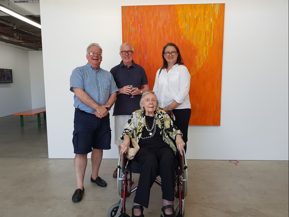 Janis Nedela, David Forrest and Lee Kinsella stand behind Carol Rudyard, in a wheelchair, with orange painting by Carol on wall behind